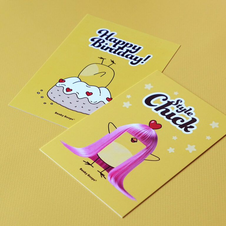 Zwei Postkarten liegen nebeneinander: Ein Küken steckt kopfüber in einem Gugelhupf, das andere Motiv zeigt ein Küken mit pinker Langhaarperücke, Produkte der Marke Beaky Boops, Küken mit Herz